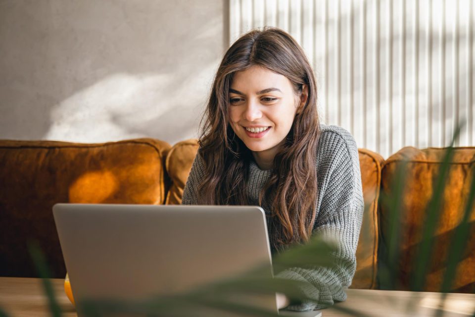 Woman with long hair sits on a brown couch using a laptop at a wooden table in a cozy room.
